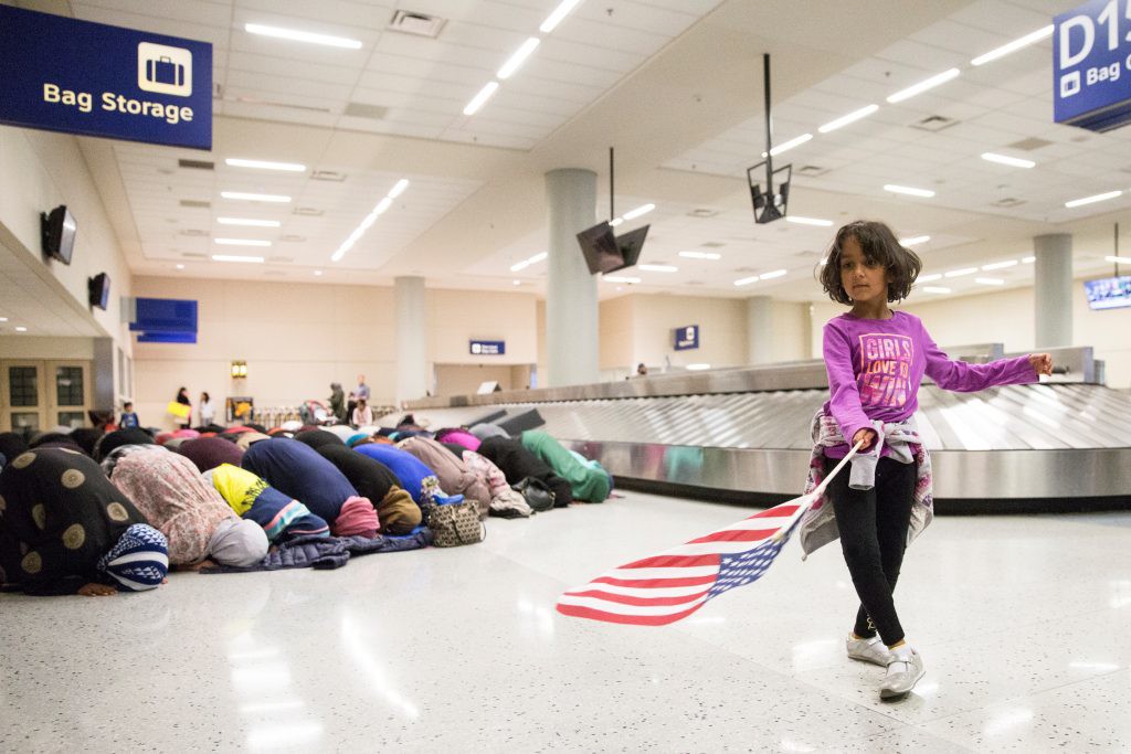 A young girl dances with an American flag while women pray behind her during a protest against the temporary travel ban